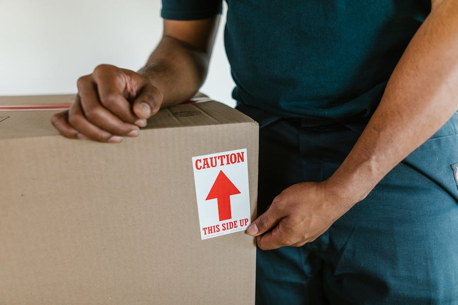 Close-up view of a person in a dark blue uniform preparing for a home relocation, holding a large cardboard box marked with a red and white caution label that reads 'CAUTION THIS SIDE UP'. The person is grasping the box firmly, with their right hand on the side of the box and their left hand on the top, indicating an active packing or loading process. In the background, the edge of a doorway and part of an interior wall are visible, suggesting the scene takes place inside a property. The box is made of brown cardboard, sealed with packing tape, and appears to contain household items. The attention to careful handling and clear signage is consistent with professional furniture transport and packing procedures, as performed by Removal Van Ilford's removals services, emphasizing safe loading practices during the moving process.