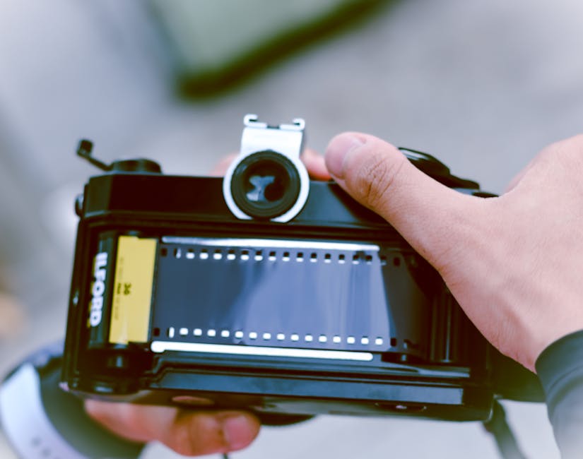 A person's hand holding a vintage film camera with a film reel visible inside the case, positioned outdoors on a paved surface. The camera features a metallic lens and a yellow film canister labeled 'EKTAN.' The background is blurred, suggesting an outdoor setting at daylight, with natural lighting illuminating the scene. The image captures the process of preparing or inspecting film for photography, aligning with careful handling associated with packing and moving equipment, as seen in home relocation or furniture transport services provided by Removal Van Ilford. The focus on the camera emphasizes precision and attention to detail characteristic of professional removals and transport logistics.