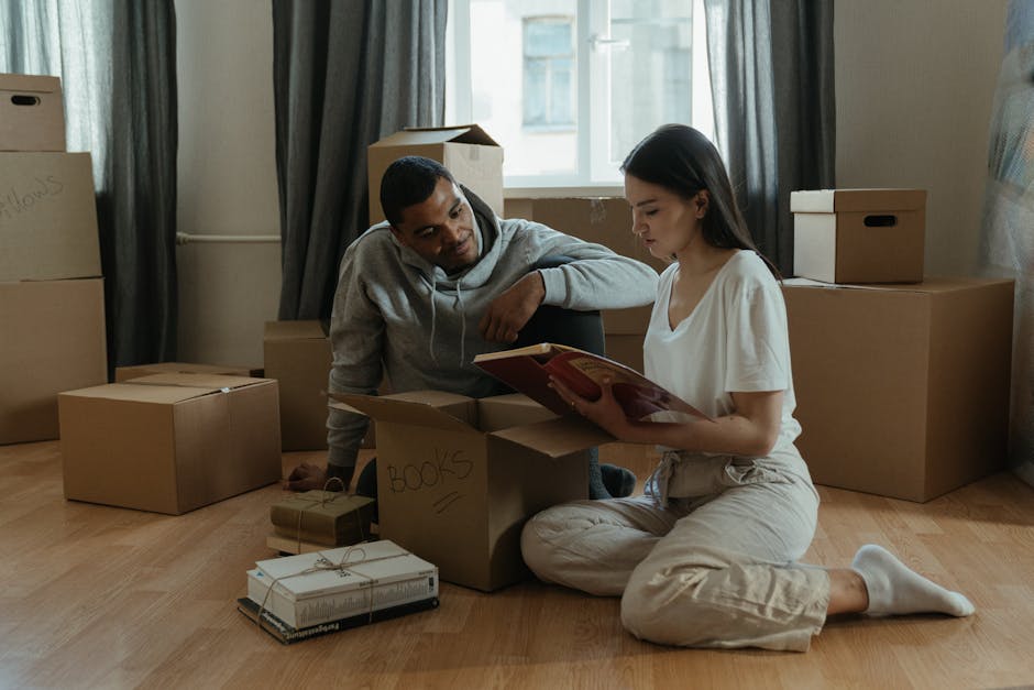 Image shows a young woman and a man in a room with wooden flooring, surrounded by numerous cardboard moving boxes, some labeled with handwritten notes such as 'BOOKS'. The woman is seated on the floor, holding a red hardcover book, while the man kneels beside her, leaning on a box, and appears to be engaging in conversation or giving instructions. The room is lit with natural light coming through a window with curtains, and additional boxes are stacked against the walls, indicating an active packing or home relocation process. The scene reflects a typical bedding and packing stage during furniture transport and moving preparations, with various materials like cardboard and packing tape visible, emphasizing the importance of proper packing and load organization in house removals. Removal Van Ilford, known for professional removals, often assists clients with similar home relocation setups, ensuring safe and efficient furniture transport.