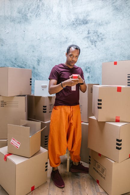 A man with medium brown skin, wearing a maroon t-shirt and bright orange trousers, is standing indoors amidst a large stack of cardboard boxes of various sizes, some sealed with red tape and others open revealing packing materials. He is holding a smartphone, appearing to review details or communicate during a home relocation process. The background features a textured, light blue and grey wall, and the floor is wooden. The scene depicts a packing or moving stage related to furniture transport and logistics, illustrating a typical aspect of house removals. Removal Van Ilford is involved in organizing and managing this packing and loading process with a focus on efficiency and care.