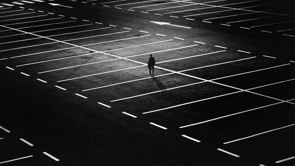 A solitary figure stands in the middle of an empty, dimly lit parking lot at night, surrounded by numerous white parking lines that create a grid pattern on the dark asphalt surface. The person is casting a long shadow due to a single overhead light source, and the overall scene emphasizes isolation and minimalism. The parking lot appears to be outside a building, with no visible vehicles or other people present. This scene may be used to illustrate challenges related to home relocation and furniture transport, such as navigating difficult staircases during large removals, as discussed on the page about common problems with Ilford staircases for large removals on removalvanilford.co.uk.