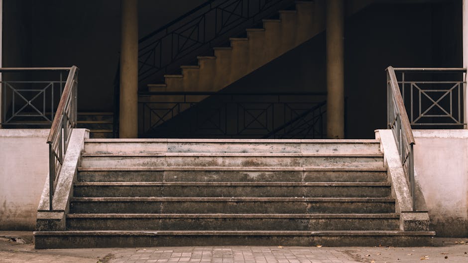 A set of concrete stairs leading up to an internal staircase within a building, with metal railings on both sides of the staircase. The steps are stained and appear weathered, showing signs of wear typical of frequent use. The interior of the building is dimly lit, with a dark staircase visible at the top, and the surrounding walls are smooth and painted in neutral tones. The external area includes a cobbled pavement at the base of the stairs, and the overall environment suggests a typical urban setting for home relocation or furniture transport. The scene reflects the challenges of navigating large items, such as furniture or boxes, through staircase layouts during a house move, which Removal Van Ilford's services are equipped to manage efficiently.