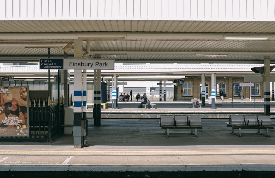 The image shows Finsbury Park railway station platform, covered by a large metal roof with evenly spaced support columns. Several wooden benches are situated along the platform, with some passengers seated, while others are walking or waiting. A black metal fence with a poster for 'Romeo and Juliet' separates the platform from the tracks. Above, a white signboard displays the station name 'Finsbury Park' in black letters, along with directional information for lifts and underground services. The station building, visible in the background, features brick walls and multiple arched windows. The scene is lit with natural daylight, with shadows cast on the platform. This setting reflects a typical urban station environment, suitable for viewing the context of home relocation or moving logistics near Ilford, as discussed on removalvanilford.co.uk, where professional removals and furniture transport services are coordinated around busy transportation hubs.