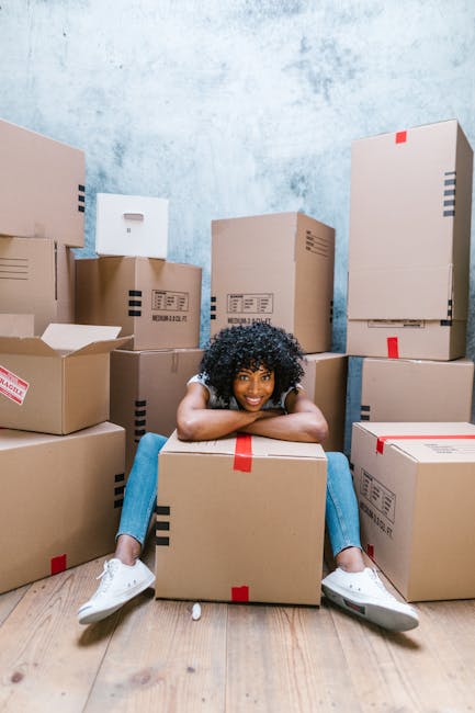 A young woman with curly black hair is sitting cross-legged on a wooden floor inside a room, surrounded by numerous brown cardboard boxes of various sizes, some sealed with red tape and others open with flaps folded. She is smiling and resting her chin on her arms, which are placed on a box in front of her. The boxes are stacked against a light blue textured wall, indicating an active packing process during a home relocation. Visible cardboard packaging materials and some empty boxes suggest the packing stage of a house move. The scene captures the preparatory steps of furniture transport and moving logistics, with the environment illuminated by natural or soft lighting, supporting the theme of professional removals services offered by Removal Van Ilford, especially for moves near IG1, ILFORD.