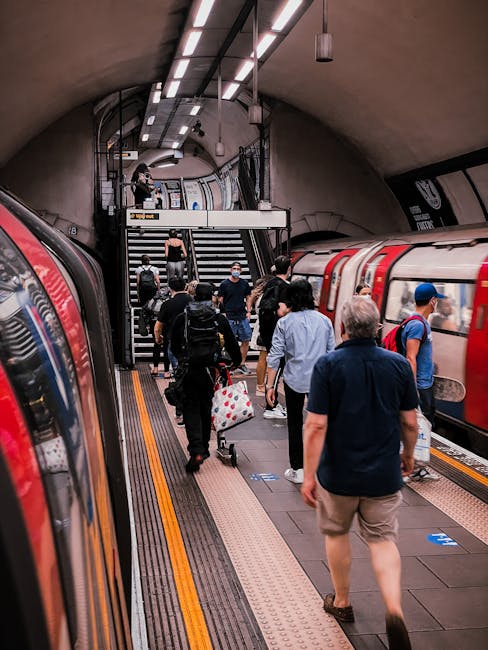 The image shows Finsbury Park railway station platform, covered by a large metal roof with evenly spaced support columns. Several wooden benches are situated along the platform, with some passengers seated, while others are walking or waiting. A black metal fence with a poster for 'Romeo and Juliet' separates the platform from the tracks. Above, a white signboard displays the station name 'Finsbury Park' in black letters, along with directional information for lifts and underground services. The station building, visible in the background, features brick walls and multiple arched windows. The scene is lit with natural daylight, with shadows cast on the platform. This setting reflects a typical urban station environment, suitable for viewing the context of home relocation or moving logistics near Ilford, as discussed on removalvanilford.co.uk, where professional removals and furniture transport services are coordinated around busy transportation hubs.