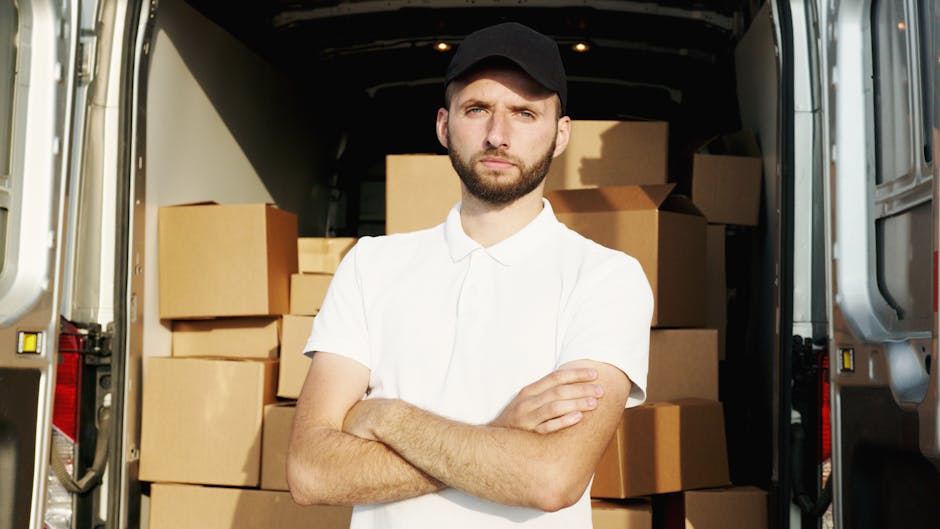 A young man with a beard and short hair, wearing a black cap and a white polo shirt, stands inside the open rear door of a moving van, which is filled with stacked cardboard boxes of various sizes. The boxes are arranged on the floor and along the sides of the van, some wrapped in plastic or packing material. The man has a serious expression, arms crossed, ready to assist with home relocation or furniture transport. The interior of the van is dimly lit, with natural light coming from outside, highlighting the packing and loading process typical of house removals services. This scene illustrates the unloading phase of a professional moving operation, with a focus on boxes and equipment suitable for packing, handling, and transporting household items, aligned with the services offered by Removal Van Ilford for urgent same-day moves in Ilford.