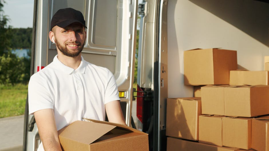 A male removal worker from Removal Van Ilford wearing a black cap and white polo shirt is loading cardboard boxes into the back of a commercial moving van. The van is parked outdoors on a clear day with green trees and a lawn visible in the background. Several brown cardboard boxes, some wrapped with plastic or packing tape, are situated inside the van, arranged in stacks near the doorway. The worker is holding one box close to his chest, preparing for a home relocation or furniture transport process. Bright natural light illuminates the scene, highlighting the details of the packaging materials, the loading area, and the transportation vehicle used by the removals company to facilitate quick and efficient packing and moving services in Ilford.