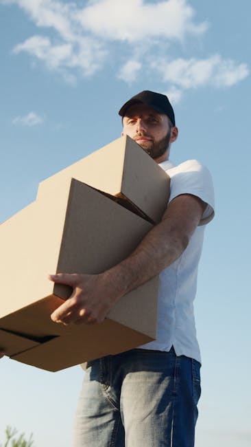 A young man with a beard and short hair, wearing a black cap and a white polo shirt, stands inside the open rear door of a moving van, which is filled with stacked cardboard boxes of various sizes. The boxes are arranged on the floor and along the sides of the van, some wrapped in plastic or packing material. The man has a serious expression, arms crossed, ready to assist with home relocation or furniture transport. The interior of the van is dimly lit, with natural light coming from outside, highlighting the packing and loading process typical of house removals services. This scene illustrates the unloading phase of a professional moving operation, with a focus on boxes and equipment suitable for packing, handling, and transporting household items, aligned with the services offered by Removal Van Ilford for urgent same-day moves in Ilford.