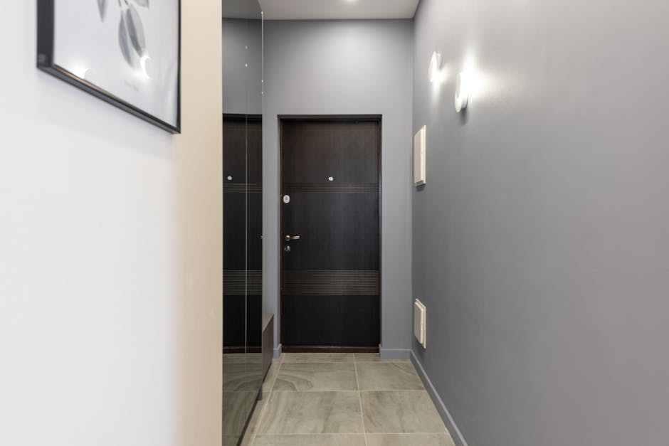 The image shows a narrow hallway inside a residential property, with a dark wooden door at the end featuring a silver door handle and a small circular peephole. To the left, there is a mirrored wardrobe with a glossy black finish, reflecting part of the corridor. The hallway walls are painted in light grey, with two white wall-mounted light fixtures providing illumination. On the right wall, there are two small rectangular vents near the floor and a white electrical box mounted higher up. The floor is tiled with large, light grey tiles arranged in a grid pattern. The space appears clean and uncluttered, suitable for a home relocation or furniture transport process, as the corridor provides access from the entrance to adjoining rooms. Removal Van Ilford may utilize this area for loading or guiding packing and moving efforts through the doorway, ensuring careful navigation of furniture and boxes in narrow hallways like this one during house removals.
