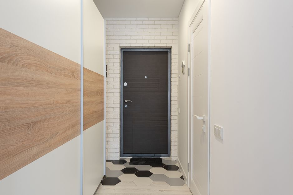 The image shows a narrow hallway inside a residential property, with a dark wooden door at the end featuring a silver door handle and a small circular peephole. To the left, there is a mirrored wardrobe with a glossy black finish, reflecting part of the corridor. The hallway walls are painted in light grey, with two white wall-mounted light fixtures providing illumination. On the right wall, there are two small rectangular vents near the floor and a white electrical box mounted higher up. The floor is tiled with large, light grey tiles arranged in a grid pattern. The space appears clean and uncluttered, suitable for a home relocation or furniture transport process, as the corridor provides access from the entrance to adjoining rooms. Removal Van Ilford may utilize this area for loading or guiding packing and moving efforts through the doorway, ensuring careful navigation of furniture and boxes in narrow hallways like this one during house removals.