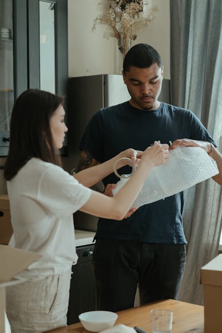 A woman and a man are inside a residential property, involved in a home relocation process. The woman, dressed in a white top, is carefully wrapping a small object in white packing paper or bubble wrap, preparing it for transportation. The man, wearing a dark T-shirt, is holding a delicate item, possibly an ornament or fragile household object, which he is possibly inspecting or preparing to pack. They are standing near a kitchen counter or dining table, with some small bowls and glasses visible on the surface. In the background, there are grey curtains partially drawn, and a tall cabinet or appliance is seen behind them. The environment is well-lit, with natural light coming through a window or door. This scene illustrates the packing phase of a comprehensive house removal service, such as those offered by Removal Van Ilford, emphasizing careful handling and packing of fragile items as part of furniture transport and moving logistics.