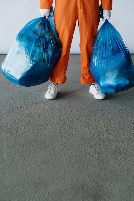 A person standing indoors on a gray carpeted floor, holding two large blue plastic recycling bags filled with waste or discarded items, with their hands covered in gray gloves. They are wearing orange
