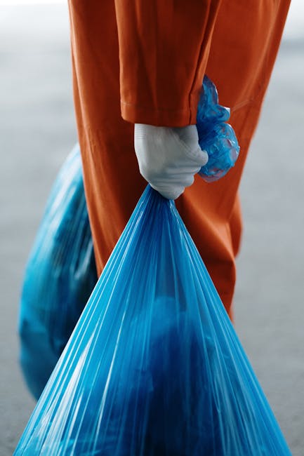 A person standing indoors on a gray carpeted floor, holding two large blue plastic recycling bags filled with waste or discarded items, with their hands covered in gray gloves. They are wearing orange